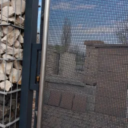 Nowoczesna brama z metalowej siatki o drobnych oczkach i pionowym, stalowym uchwytem, przylegająca do gabionu wypełnionego jasnymi kamieniami. Widok na fragment budynku i błękitne niebo przez siatkę.