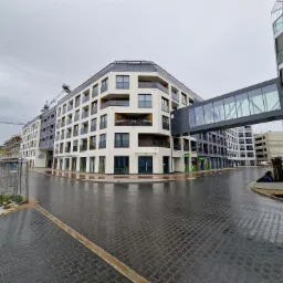 Nowoczesny budynek mieszkalny z białymi elewacjami i szklanym łącznikiem, widok na mokry chodnik z kostki brukowej po deszczu, w tle plac budowy z dźwigiem.