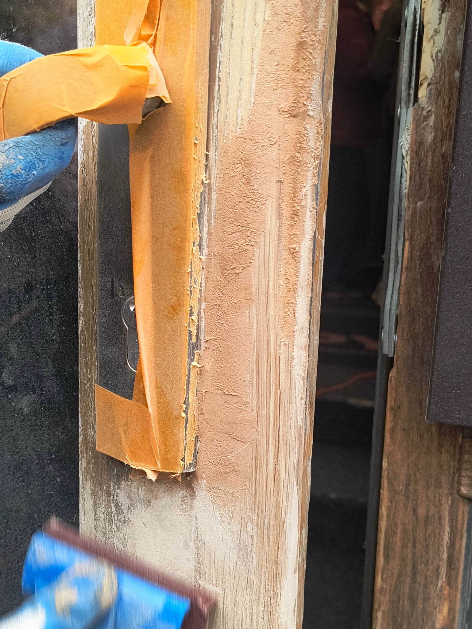 Szlifowanie szpachli na ościeżnicy drzwiowej, zabezpieczonej taśmą malarską. Widoczne ślady po szpachlowaniu i narzędzie w dłoni w rękawicy ochronnej.