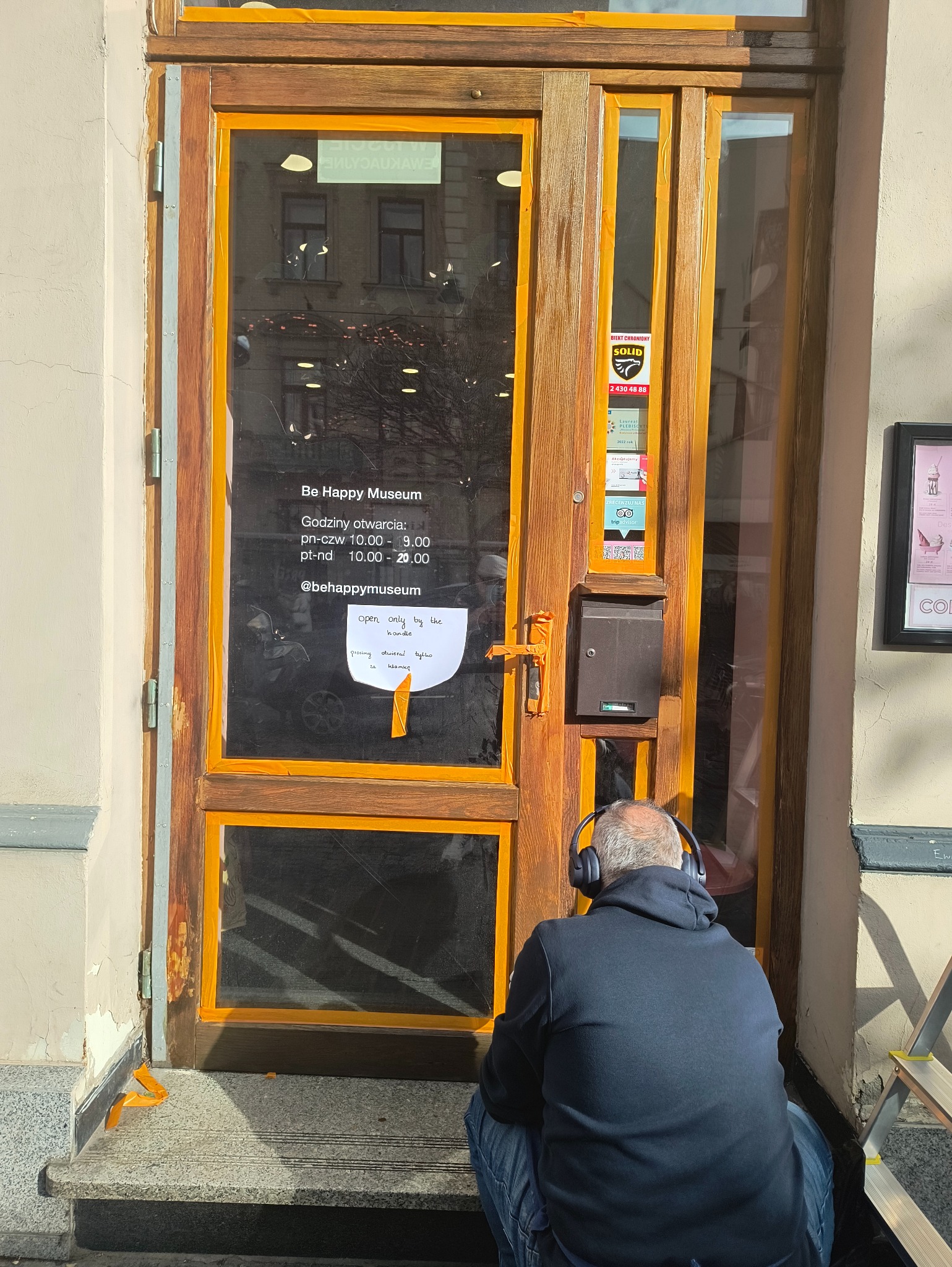 Renowacja drewnianych drzwi wejściowych do muzeum, zabezpieczonych taśmą malarską. Mężczyzna w słuchawkach siedzi na schodach, obok drabina.