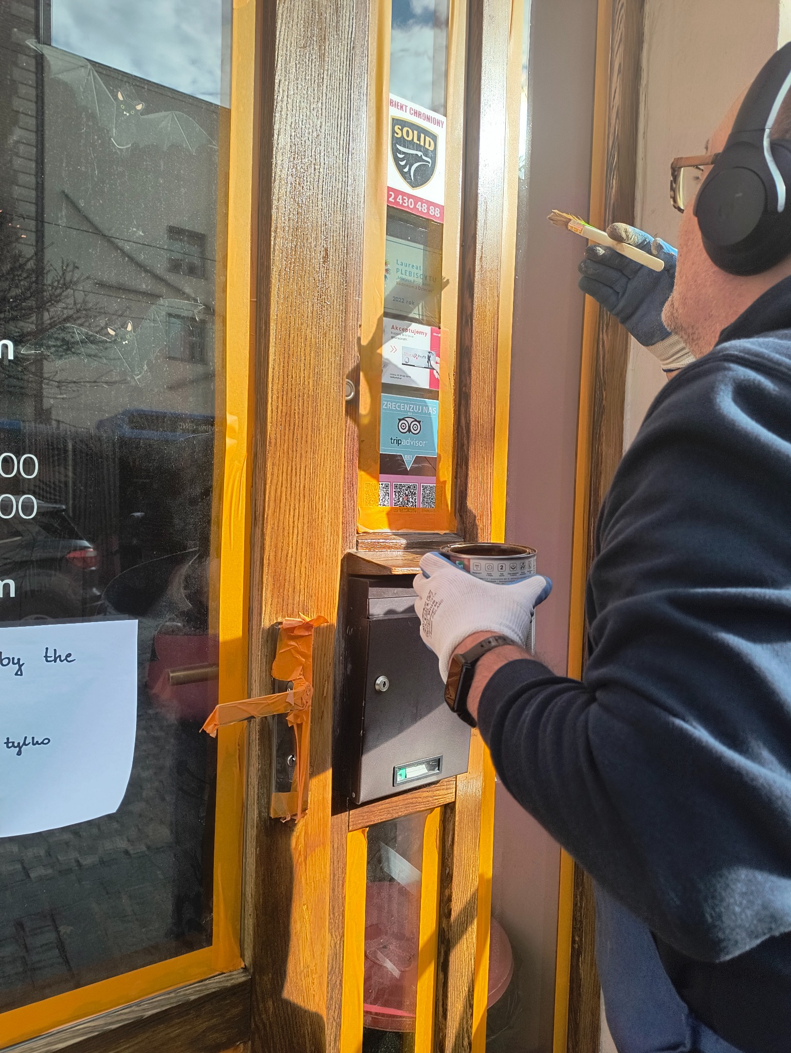 Mężczyzna w słuchawkach maluje drewniane ramy okienne zabezpieczone taśmą malarską. Widoczna puszka z farbą i pędzel. Na szybie naklejki i ogłoszenia.