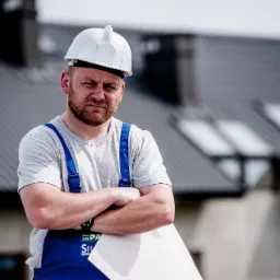 Zamyślony dekarz w białym kasku i niebieskim kombinezonie, z planami w ręku, na tle domu z ciemnym dachem z blachy trapezowej.