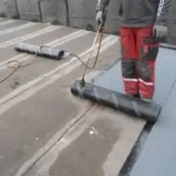 Pracownik w czerwonych spodniach z nakolannikami, używa palnika do zgrzewania papy na płaskim dachu, widoczne pasy zgrzanego materiału i rolka papy do ułożenia.