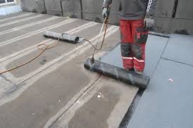 Pracownik w czerwonych spodniach z nakolannikami, używa palnika do zgrzewania papy na płaskim dachu, widoczne pasy zgrzanego materiału i rolka papy do ułożenia.