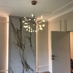 Nowoczesne wnętrze z dekoracyjną ścianą imitującą marmur, oświetloną listwami LED, oraz designerską lampą sufitową w kształcie rozgałęzionego drzewa z białymi kulkami, widok na otwarte białe drzwi.