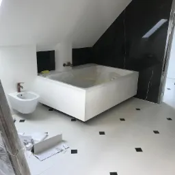 Nowoczesna łazienka w trakcie wykańczania: biała wanna obudowana płytkami, bidet z miedzianą armaturą, czarne płytki imitujące marmur na ścianie pod skosem, biała podłoga z czarnymi akcentami...