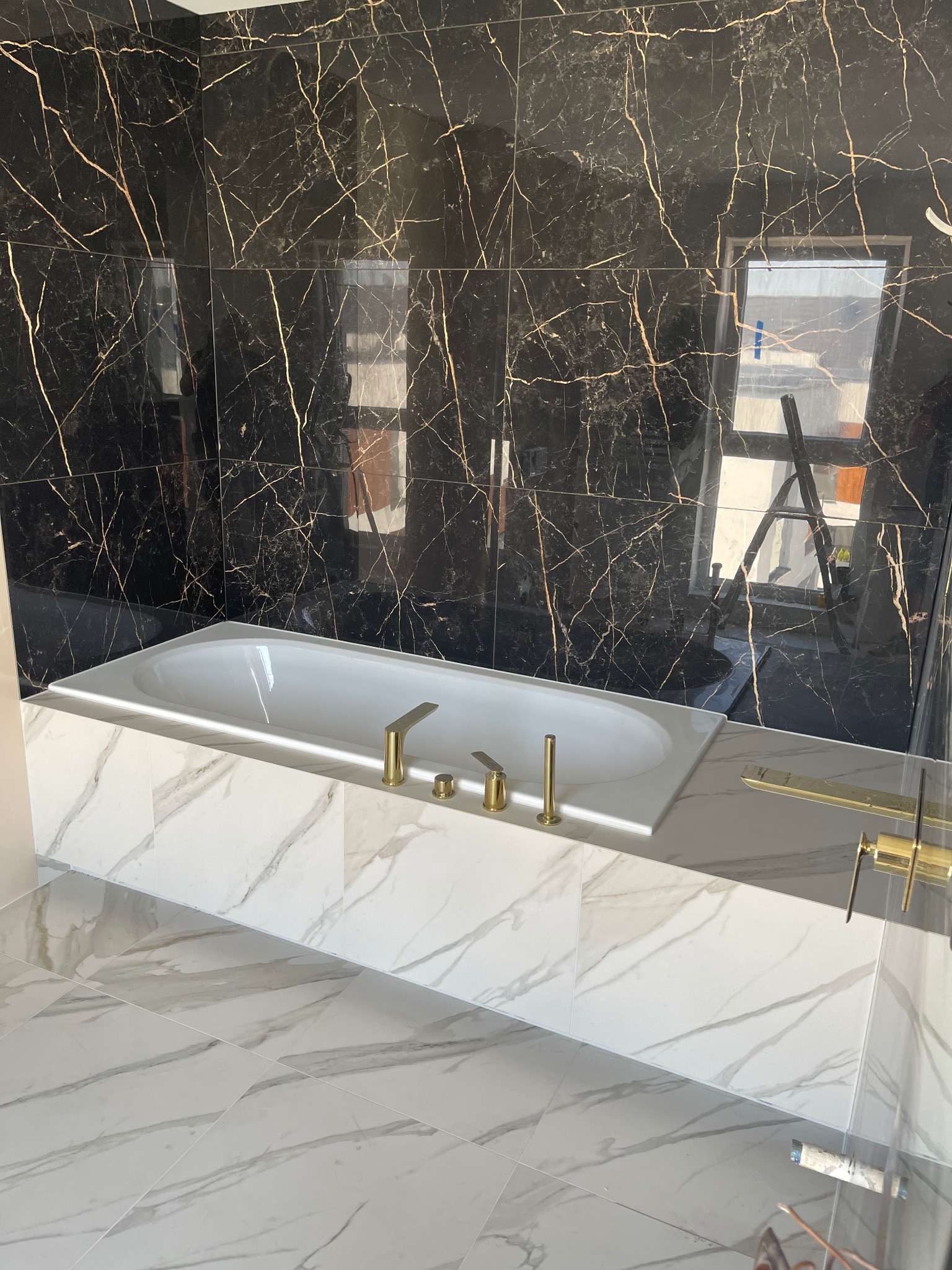 Wykończona łazienka z czarnymi płytkami imitującymi marmur na ścianach i białymi płytkami na podłodze, wanna obudowana białymi płytkami z złotą armaturą, widoczne odbicie drabiny w oknie.