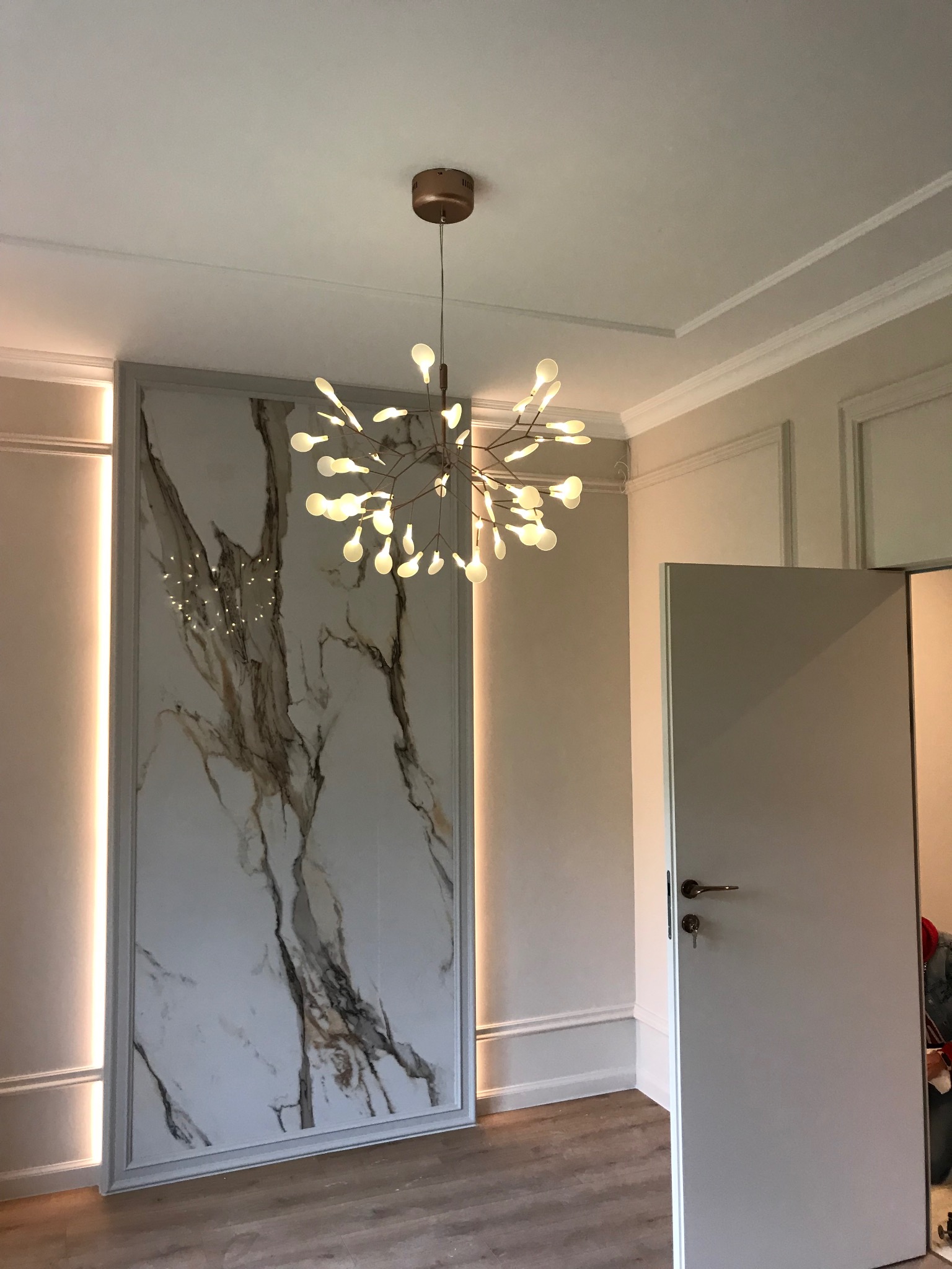 Nowoczesne wnętrze z dekoracyjną ścianą imitującą marmur, oświetloną listwami LED, oraz designerską lampą sufitową w kształcie rozgałęzionego drzewa z białymi kulkami, widok na otwarte białe drzwi.