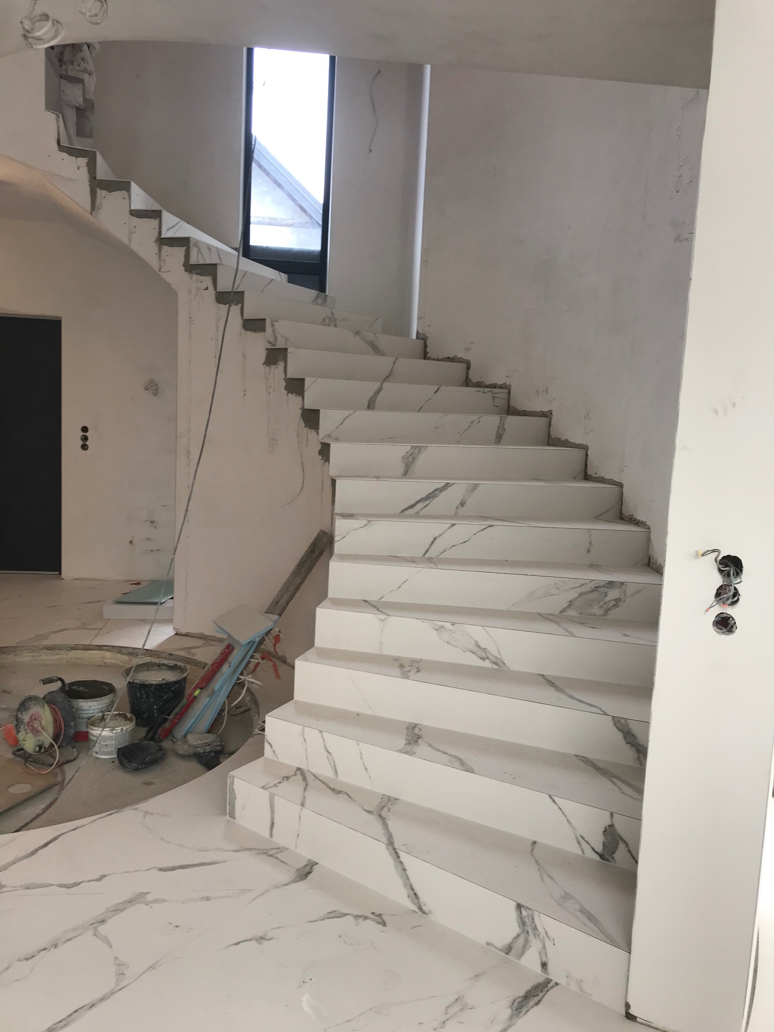Schody wewnętrzne wyłożone białymi płytkami imitującymi marmur, w trakcie prac wykończeniowych, widoczne wiaderka z zaprawą i narzędzia.