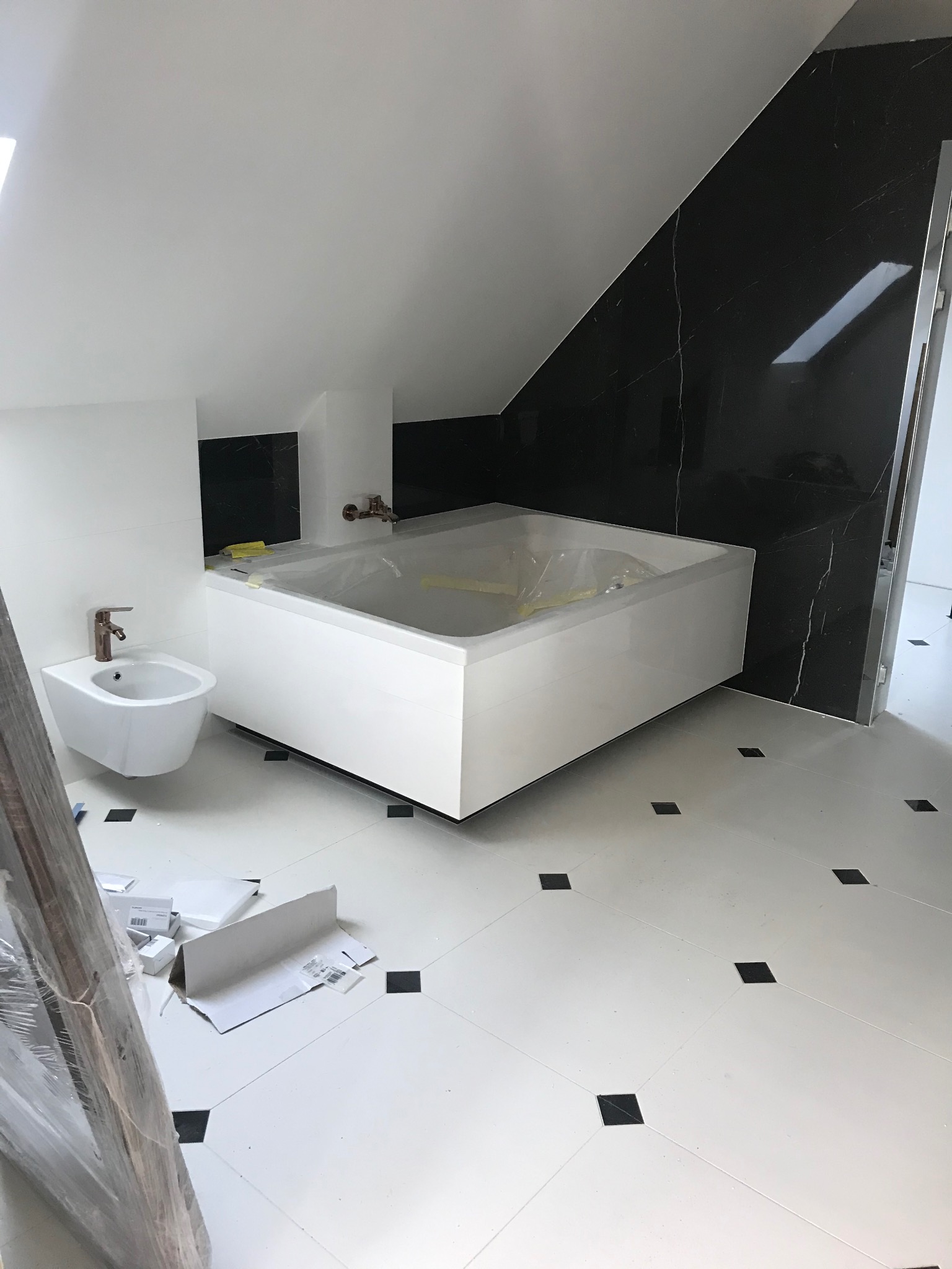 Nowoczesna łazienka w trakcie wykańczania: biała wanna obudowana płytkami, bidet z miedzianą armaturą, czarne płytki imitujące marmur na ścianie pod skosem, biała podłoga z czarnymi akcentami...