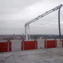 Wylewanie betonu na strop budynku z widokiem na miasto w tle, pracownicy w odblaskowych kamizelkach obsługują pompę do betonu.