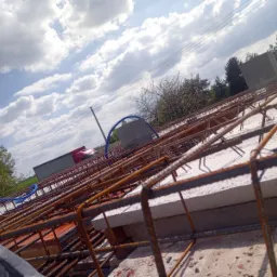 Siatka zbrojeniowa na stropie z widocznymi kształtkami betonowymi, niebieskie rury instalacyjne oraz budynek w tle, całość sfotografowana pod kątem w słoneczny dzień.