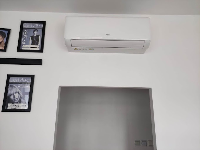 Biały klimatyzator AUX zamontowany na ścianie w salonie, obok wiszą oprawione certyfikaty fryzjerskie.