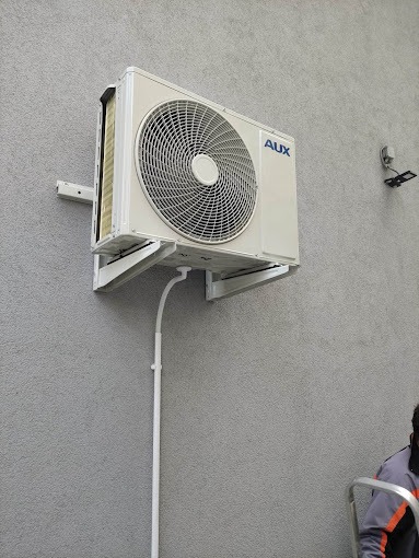 Biała jednostka zewnętrzna klimatyzatora marki AUX zamontowana na szarej, teksturowanej ścianie budynku, widoczne białe rurki instalacyjne i fragment drabiny z osobą w tle.