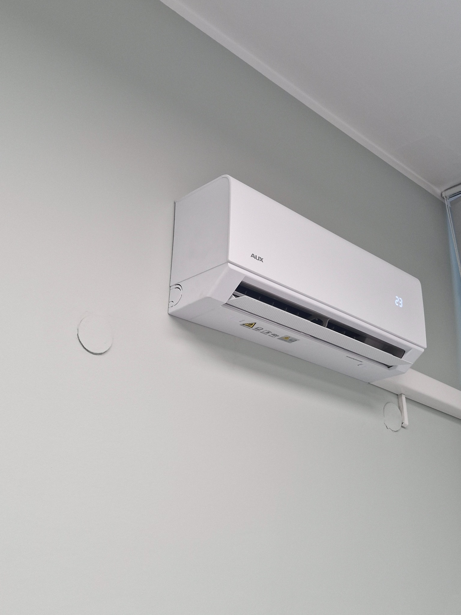 Biała klimatyzacja AUX w pomieszczeniu na jasnej ścianie. Widoczne ślady po poprzedniej instalacji, wyświetlacz temperatury wskazuje 23 stopnie.