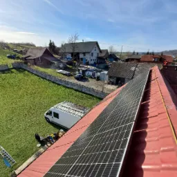 Widok z góry na fragment dachu z panelami słonecznymi w Tarnowie, z białym busem i pracownikami na trawie w tle oraz zabudowaniami wiejskimi w oddali.