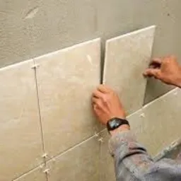 Układanie beżowych płytek ceramicznych na ścianie z użyciem krzyżyków dystansowych, widoczne dłonie w trakcie pracy i fragment zegarka na nadgarstku.