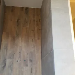 Wnętrze kabiny prysznicowej z drewnopodobną okładziną ścienną i szarą glazurą, widoczne dwa otwory w okładzinie, jasna ściana w tle.