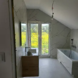 Nowoczesna łazienka na poddaszu z wanną obudowaną płytkami imitującymi marmur, drewnianą szafką pod umywalką i dużym oknem z widokiem na łąkę.