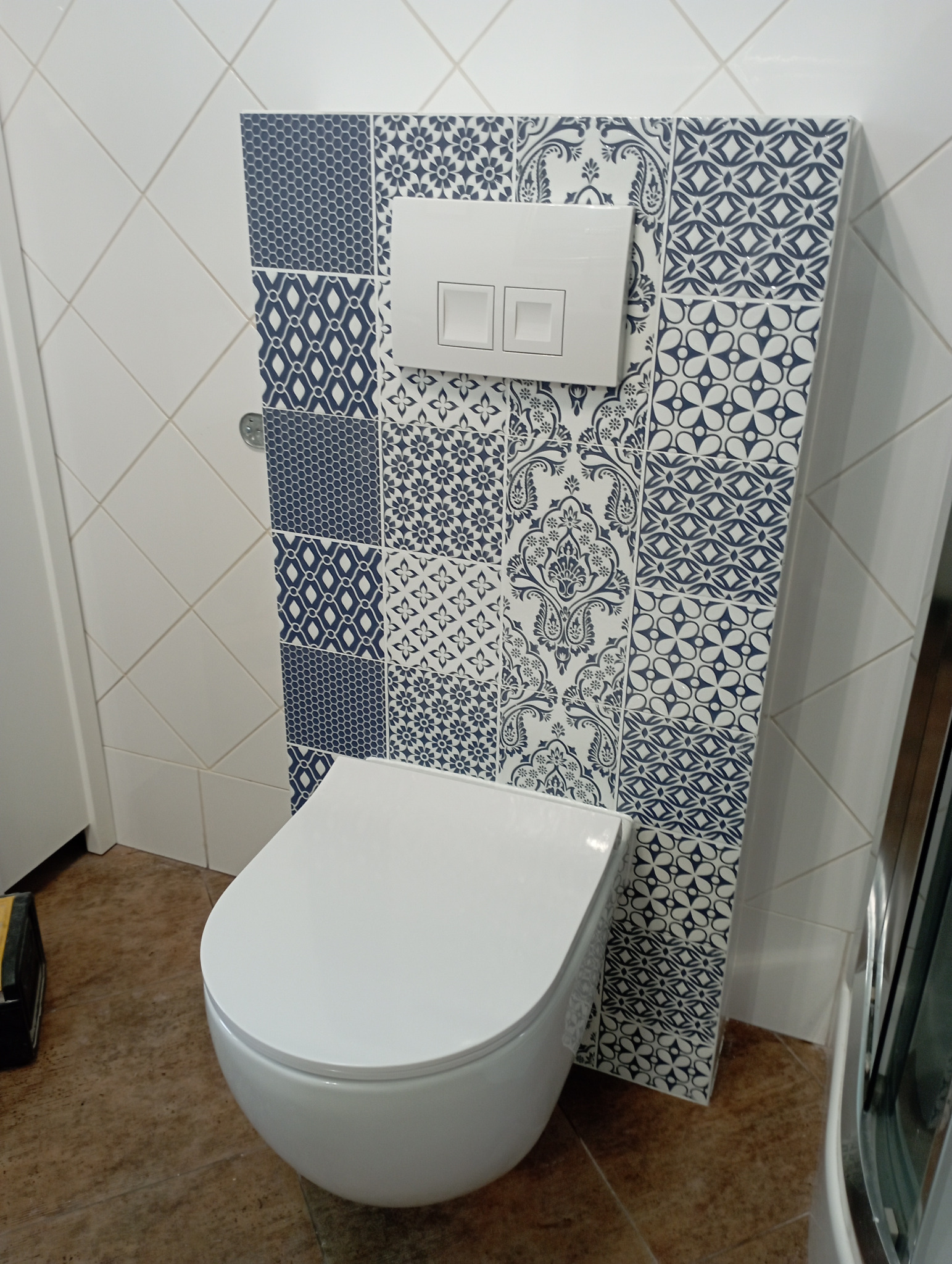 Wisząca toaleta z białą deską i przyciskiem spłukującym na ścianie wyłożonej dekoracyjnymi, niebiesko-białymi płytkami w geometryczne i kwiatowe wzory, brązowa podłoga, widoczny fragment białej...