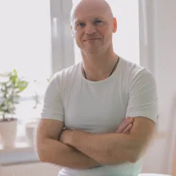 Uśmiechnięty mężczyzna w białej koszulce i sportowych spodenkach, siedzący na białym stole do masażu, z założonymi rękami, w tle okno i roślina doniczkowa.