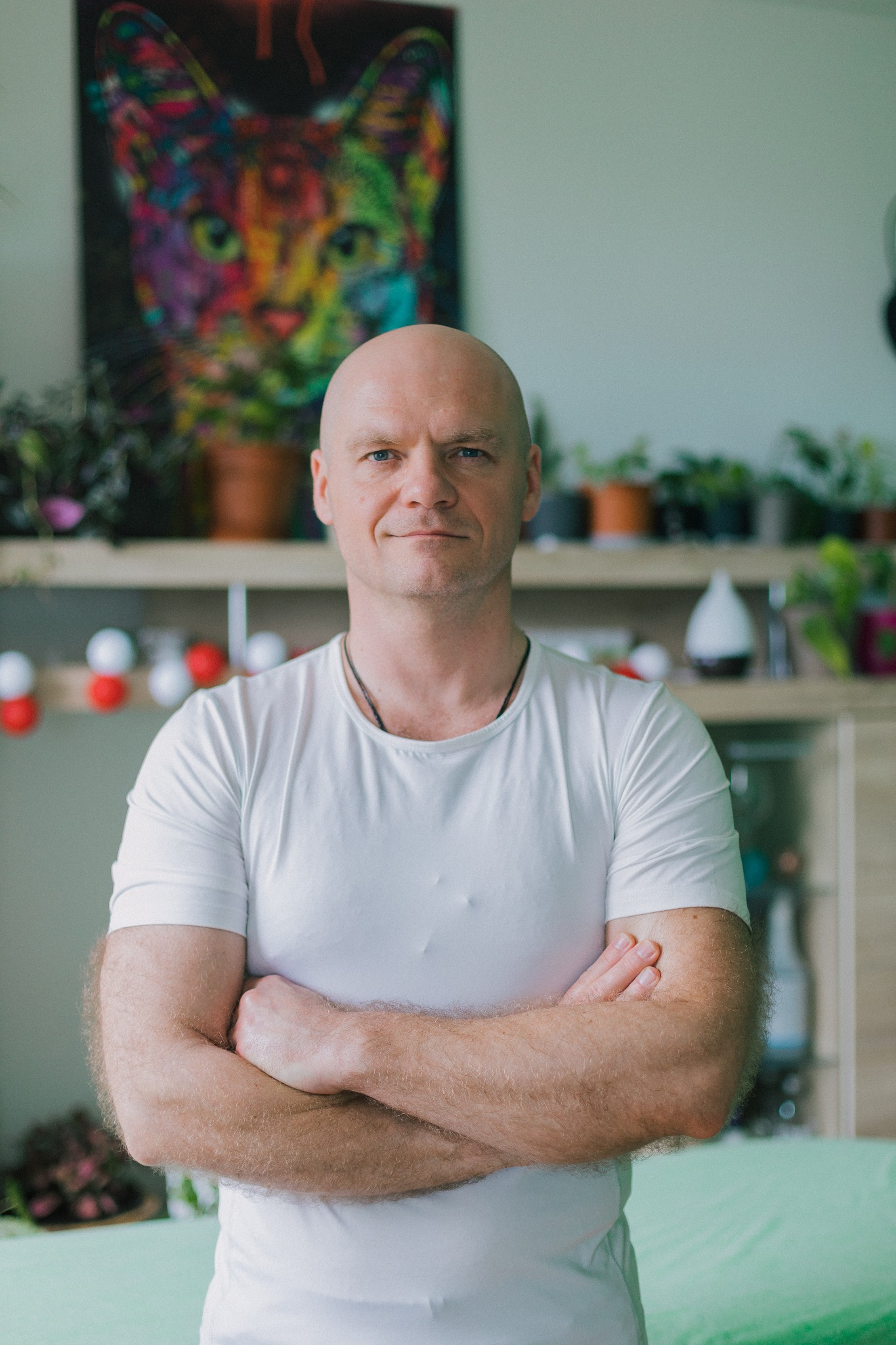 Portret mężczyzny z założonymi rękami na tle gabinetu z obrazem kota, roślinami i dyfuzorem zapachowym.