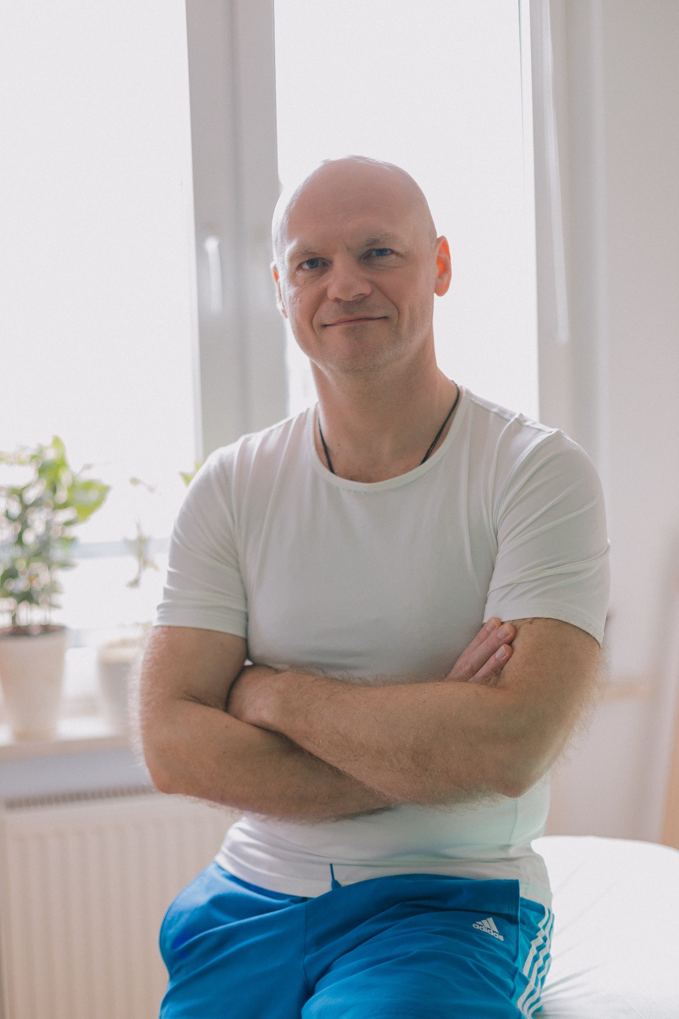 Uśmiechnięty mężczyzna w białej koszulce i sportowych spodenkach, siedzący na białym stole do masażu, z założonymi rękami, w tle okno i roślina doniczkowa.