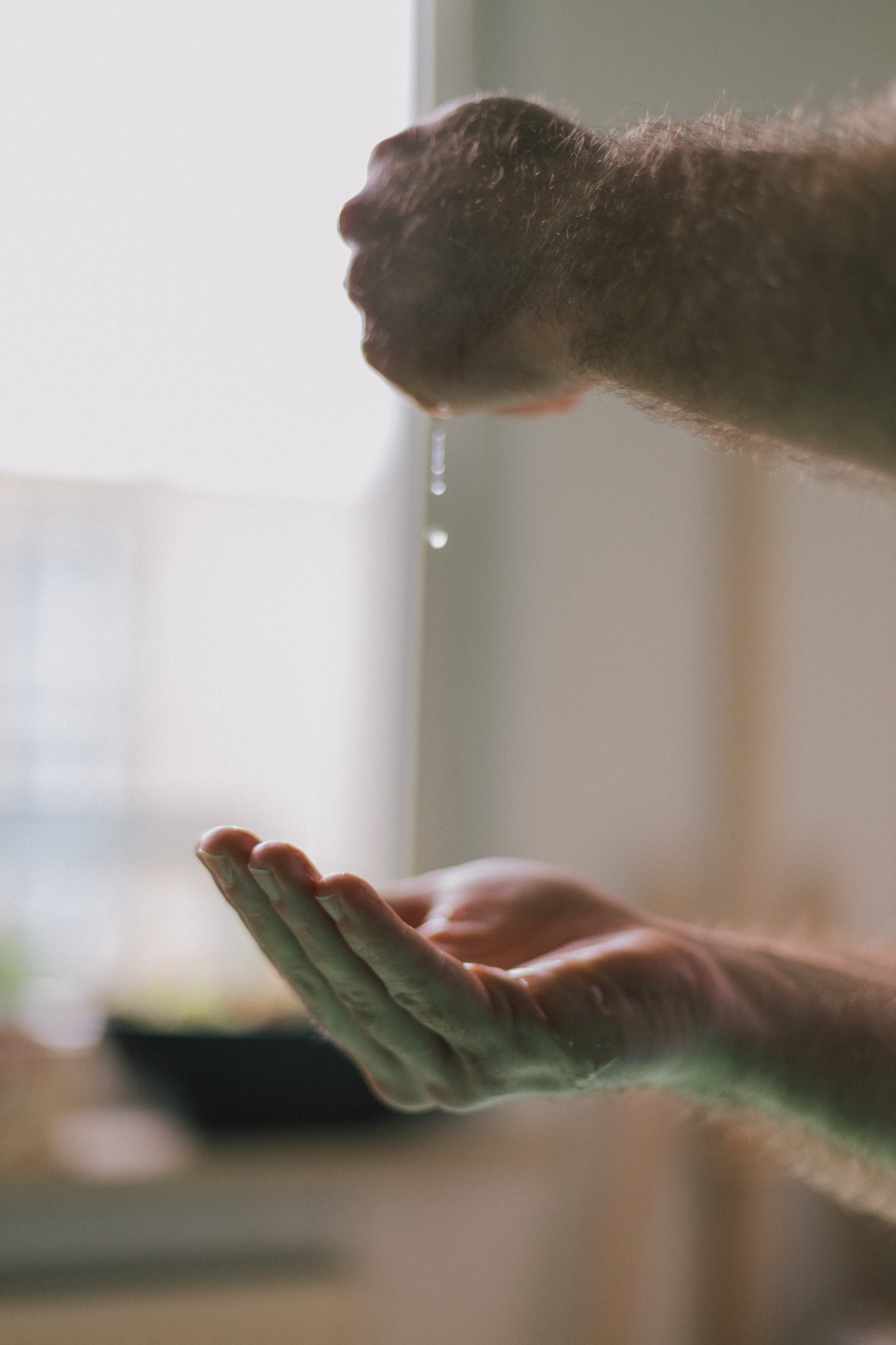 Krople cieczy wyciskane z zaciśniętej dłoni do otwartej dłoni poniżej, jasne tło okna.