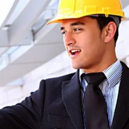 Młody mężczyzna w kasku budowlanym i garniturze, uśmiechnięty, na tle konstrukcji budynku.