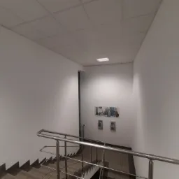 Klatka schodowa w biurowcu z pomalowanymi na biało ścianami, szarymi schodami i metalową balustradą. Na ścianie plakat z produktami i dwie małe gablotki.