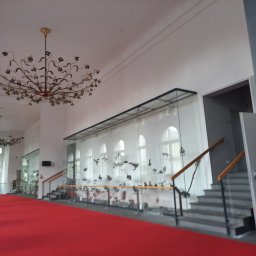 malowanieSMART - Przestronne wnętrze z czerwonym dywanem, zdobionym żyrandolem w kształcie kwiatów, szklanymi gablotami z ekspozycją oraz schodami z drewnianymi poręczami.