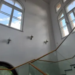 Wysokie, jasne wnętrze z białymi ścianami, oknami z widokiem na błękitne niebo, szklaną balustradą schodów i minimalistycznymi kinkietami.