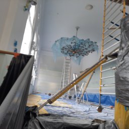 malowanieSMART - Przygotowanie do prac wykończeniowych w pomieszczeniu z żyrandolem, zabezpieczonym folią, widoczne rusztowania i folia ochronna na podłodze.