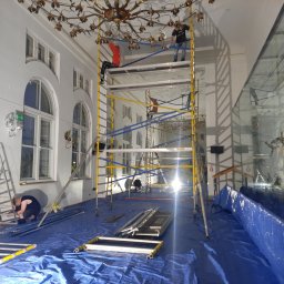 malowanieSMART - Wnętrze wysokiego pomieszczenia z marmurowymi oknami i żyrandolem w trakcie przygotowania do prac remontowych; podłoga zabezpieczona niebieską folią, rozstawione rusztowania i drabiny, dwóch...
