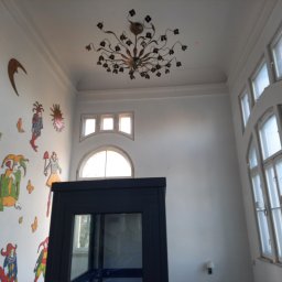 malowanieSMART - Wysokie wnętrze z dekoracjami ściennymi w postaci kolorowych postaci, ozdobnym żyrandolem i oknami z podziałami.