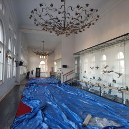 malowanieSMART - Przygotowanie do malowania w przestronnym wnętrzu z wysokimi oknami i dekoracyjnym żyrandolem, zabezpieczenie podłogi niebieską folią ochronną.