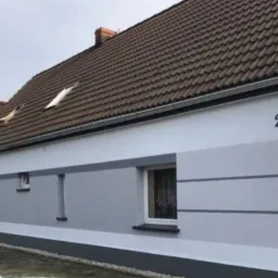 Odnowiona elewacja domu z numerem 27, pomalowana w odcieniach szarości i bieli, z widocznymi elementami wykończeniowymi i brązowym dachem.