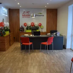 Wnętrze biura z logo Generali i Proama na ścianie, biurko z laptopem, kwiatami i tortem, czerwone krzesła, balony i dekoracje świąteczne.