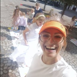 FUN-Dżasta Animator Justyna Potulicka - Uśmiechnięta kobieta w pomarańczowym daszku robi selfie na tle dzieci grających w grę na chodniku podczas imprezy plenerowej.