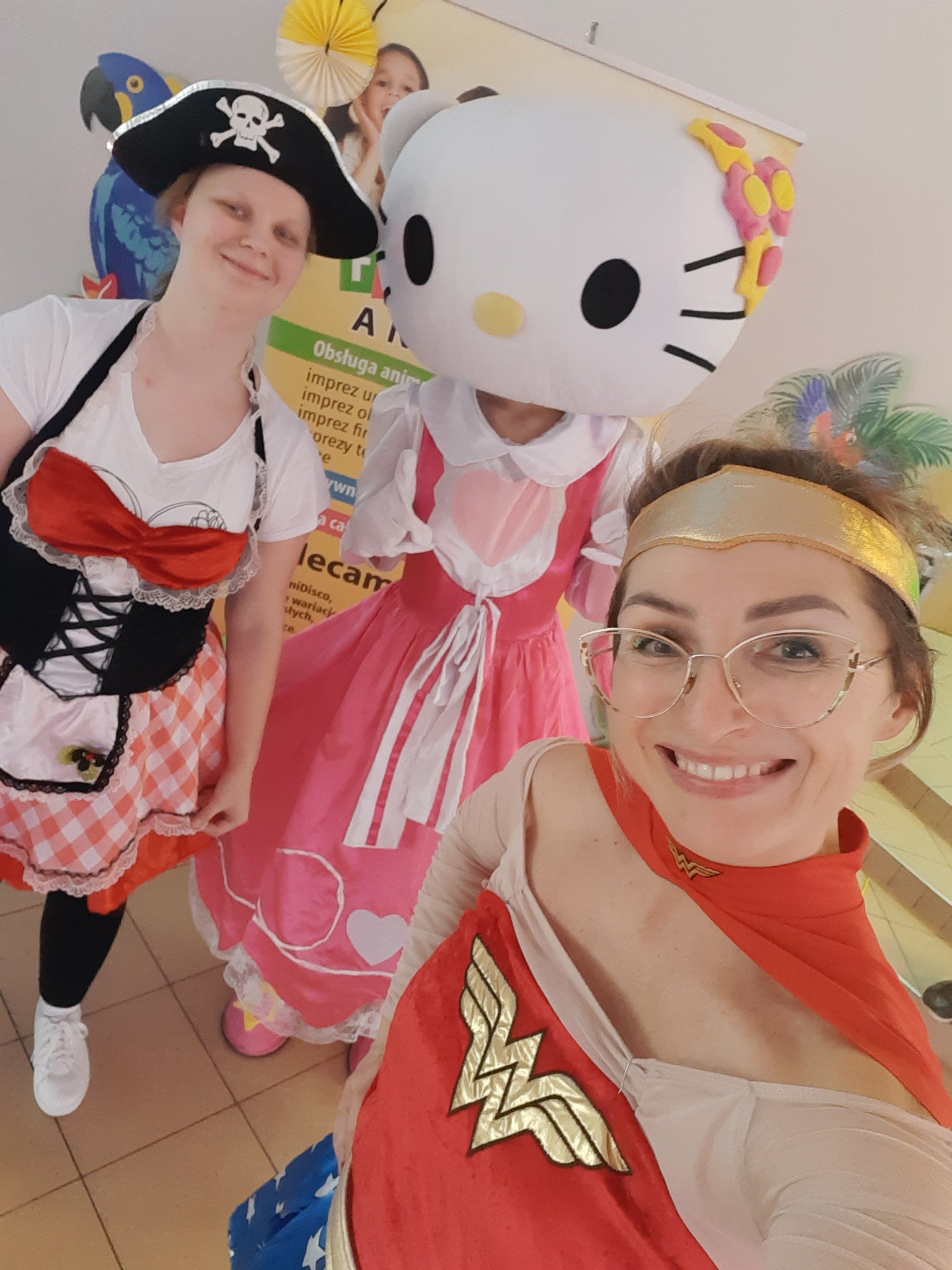 Trzy osoby w przebraniach: pirat z czaszką na kapeluszu, Hello Kitty w różowej sukience i Wonder Woman z uśmiechem na twarzy, pozują do selfie na tle plakatu.