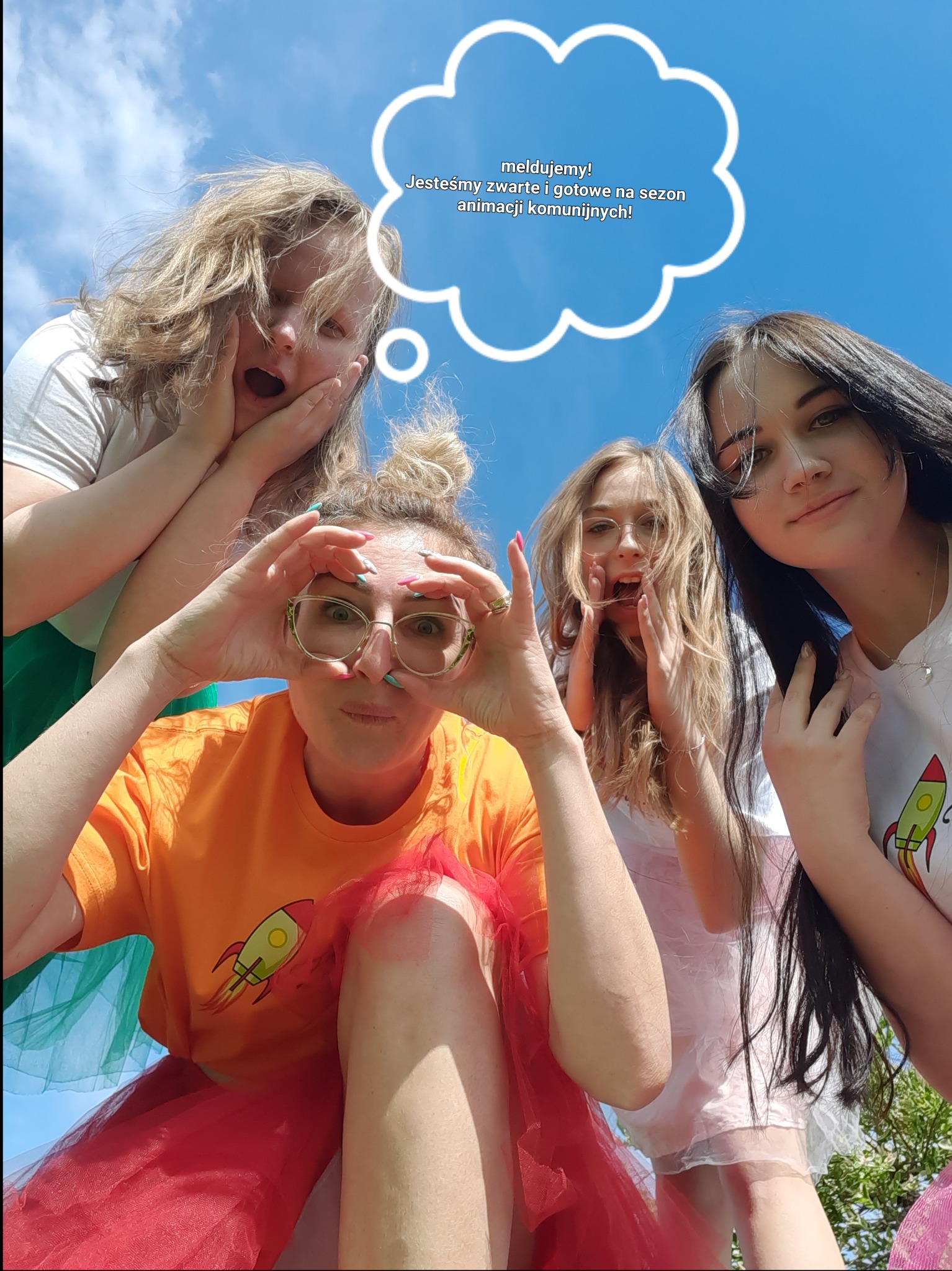 Grupa czterech młodych, uśmiechniętych kobiet w barwnych strojach, w tym jedna w pomarańczowej koszulce z rakietą i czerwonej tiulowej spódnicy, pozuje na tle błękitnego nieba z chmurką dialogową...