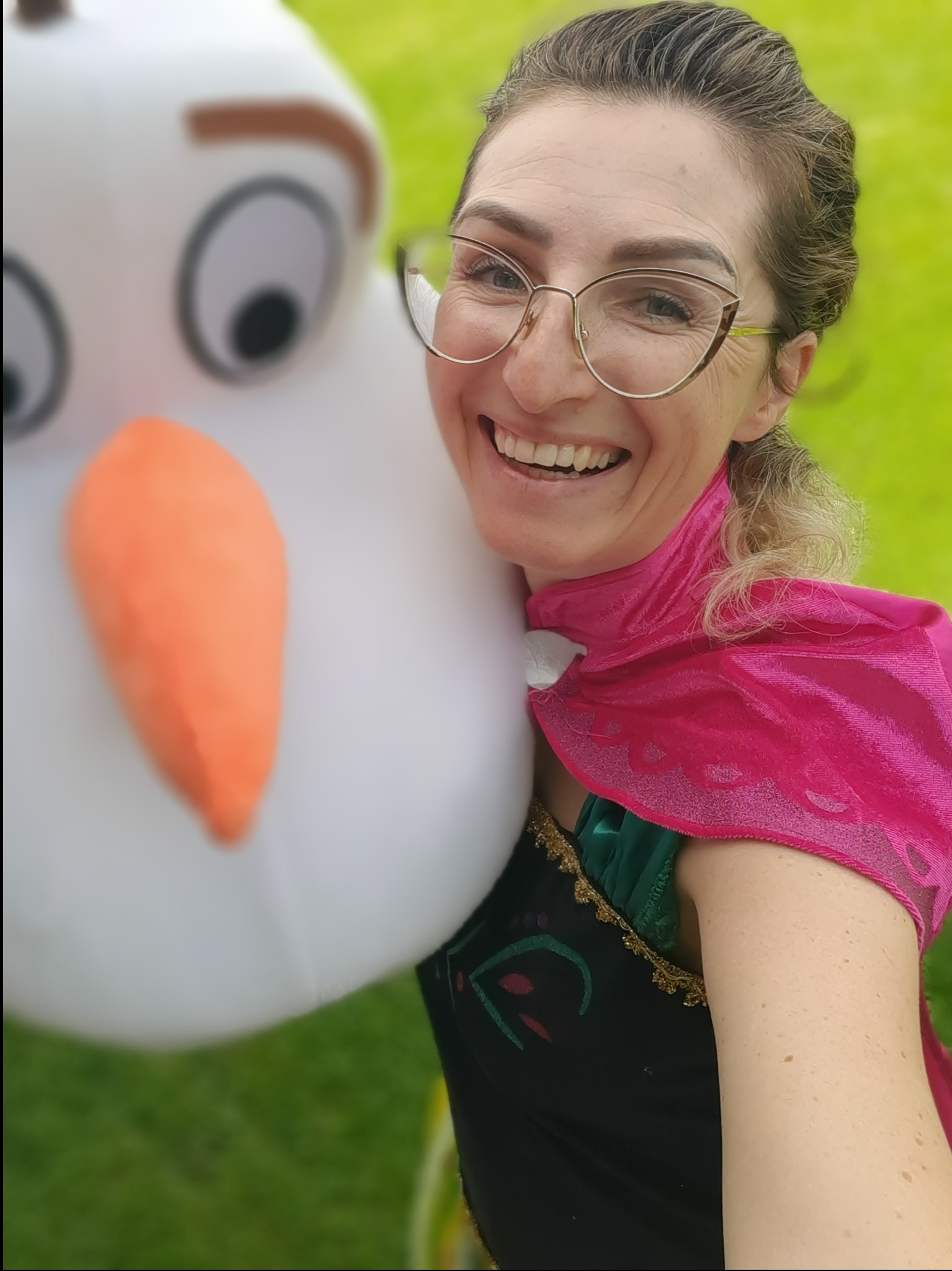 Uśmiechnięta kobieta w okularach i kostiumie Anny z 'Krainy Lodu' pozuje z dmuchanym Olafem na tle zielonej trawy.