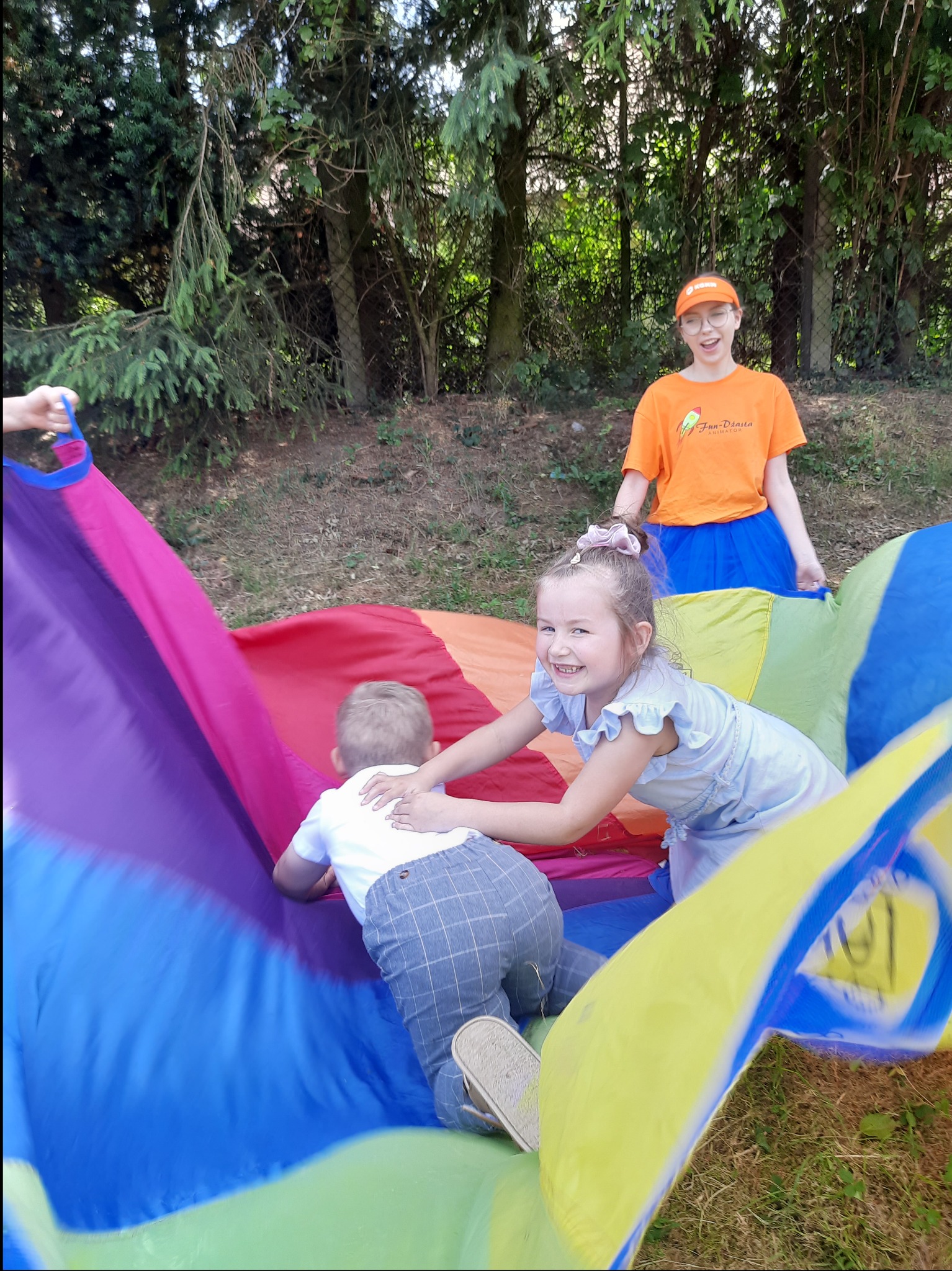 Dzieci bawią się pod kolorową chustą animacyjną, nad nimi uśmiechnięta animatorka w pomarańczowej koszulce z logo firmy.
