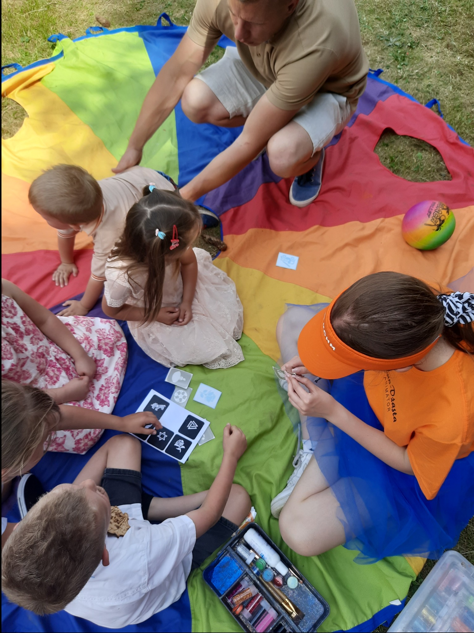 Animator dla dzieci maluje twarze podczas zabawy na kolorowej chuście animacyjnej, widoczne farbki i szablony.