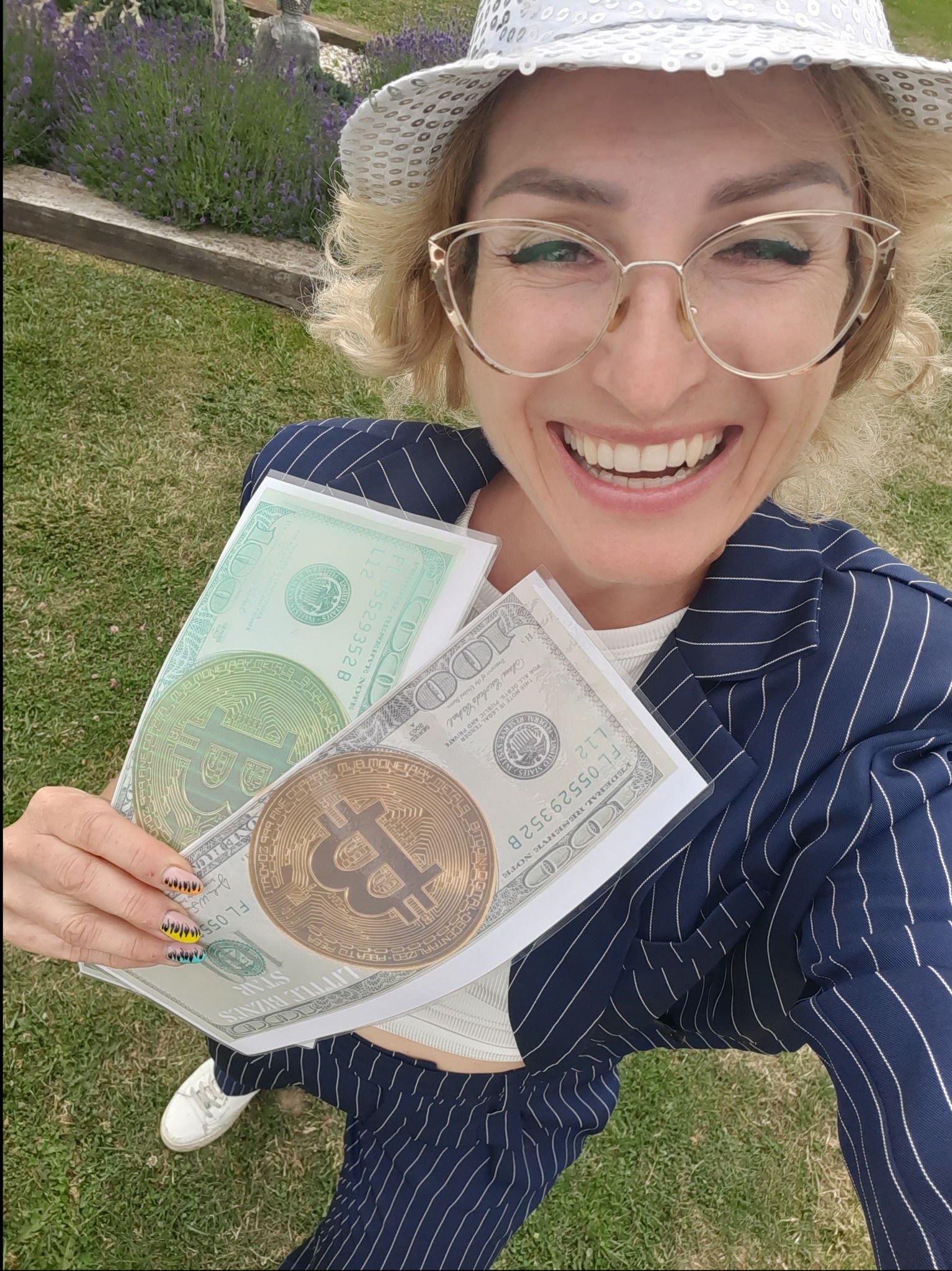 Uśmiechnięta kobieta w kapeluszu i okularach, trzymająca w rękach rekwizyty: banknoty dolarowe z wizerunkiem Bitcoin, na tle trawy i lawendy.