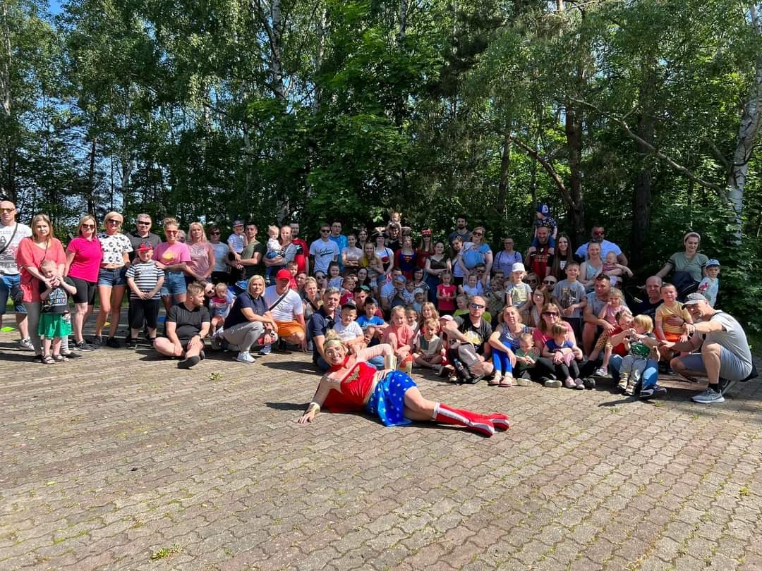 Grupowe zdjęcie dużej grupy osób, w tym dzieci, z osobą przebraną za superbohatera w kostiumie leżącą na pierwszym planie, na tle drzew i brukowanej powierzchni.