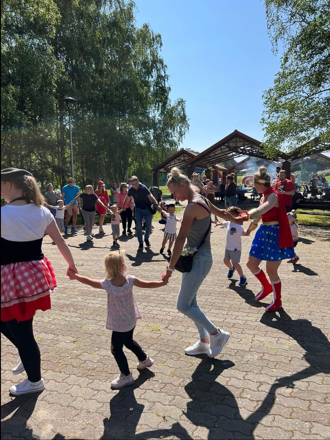 Grupa dzieci i dorosłych trzymających się za ręce, tworzących koło na placu z kostki brukowej. Wśród nich osoba przebrana za superbohaterkę w kostiumie z gwiazdami i czerwonych butach. W tle...