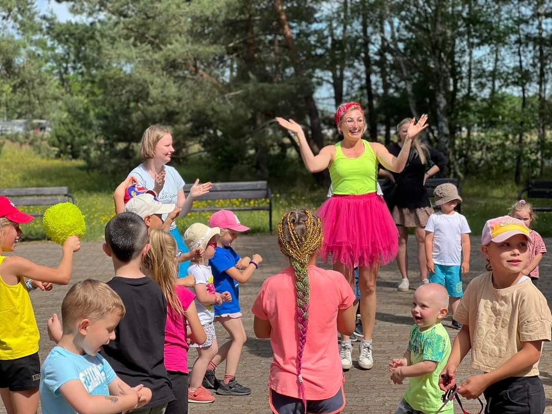 Kobieta w neonowej koszulce i różowej tutu prowadzi grupę dzieci w aktywności na świeżym powietrzu, na tle zieleni i ławek parkowych.