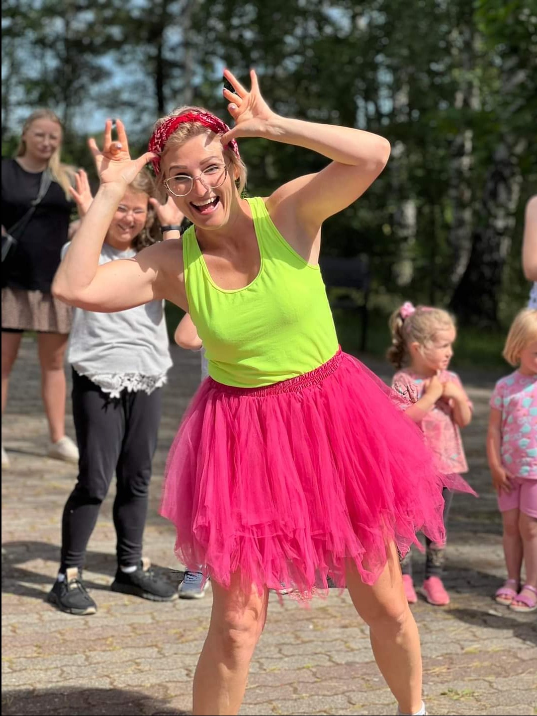 Animator w neonowej koszulce i różowej tiulowej spódnicy, robiąca śmieszną minę z uniesionymi rękami, otoczona przez dzieci podczas animacji na zewnątrz.