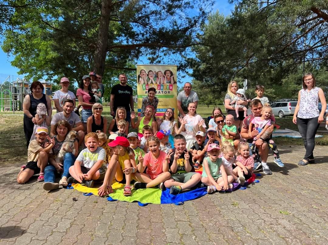 Duża grupa dzieci i dorosłych pozuje do zdjęcia na zewnątrz, na kolorowej płachcie, za nimi widoczny baner z napisem 'Fundusz Animator' i drzewa.